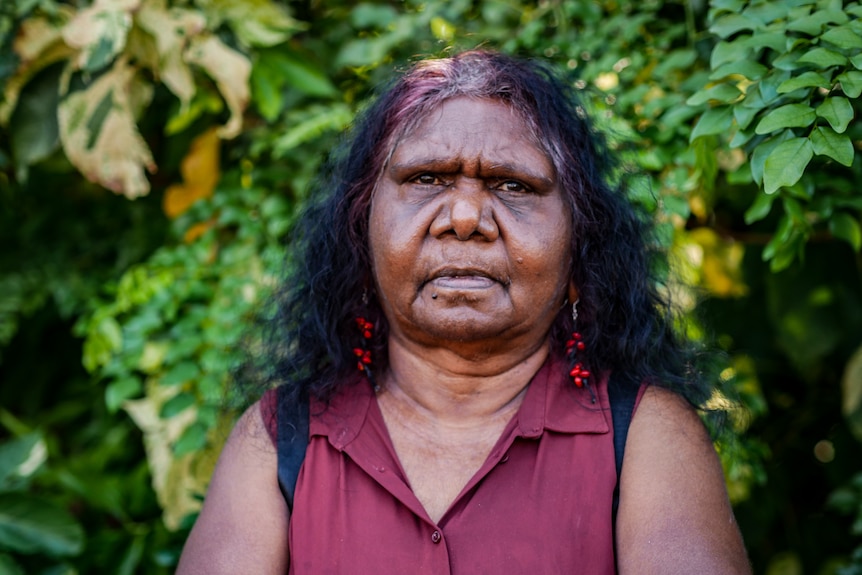 A middle aged woman wears a purple shirt and looks sternly at the camera. 