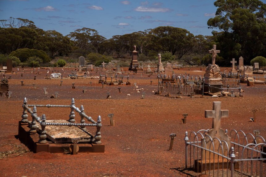 A bare red dirt graveyard with old crosses and some metal fences with trees in the background