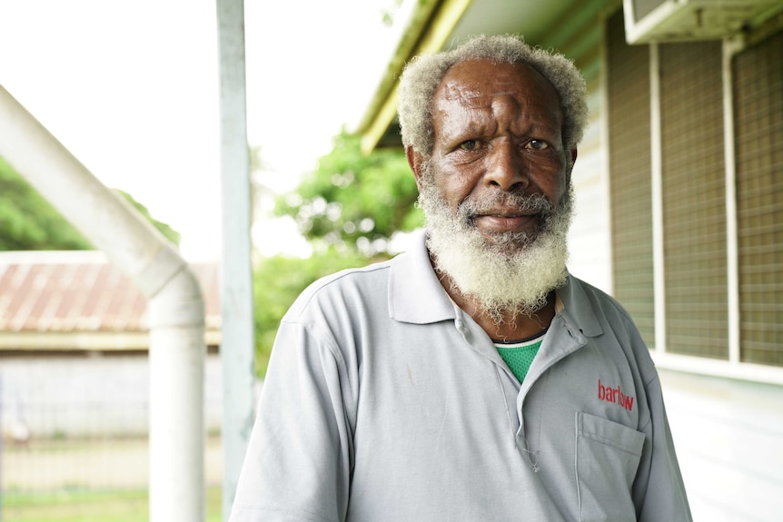 An older black man with a grey beard wearing a grey shirt stands outside a building.