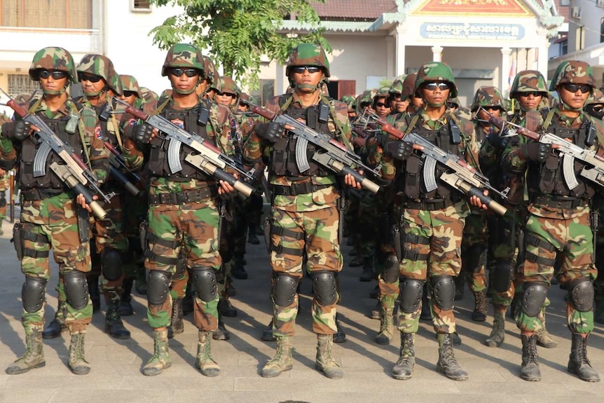 Cambodian men in army fatigues stand to attention holding rifles.