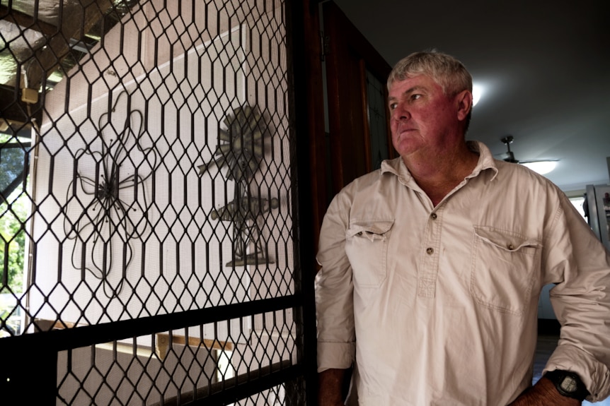 a man in a cotton long sleeved shirt stands in the door way of a home and looks out through the open screen door