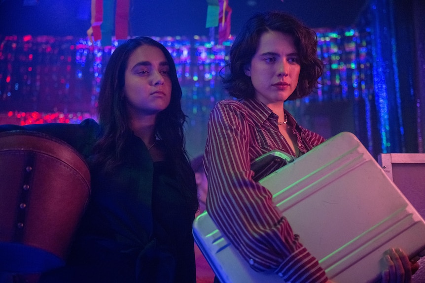A film still of Geraldine Viswanathan and Margaret Qualley in a nightclub. Qualley is holding a briefcase under her arm.