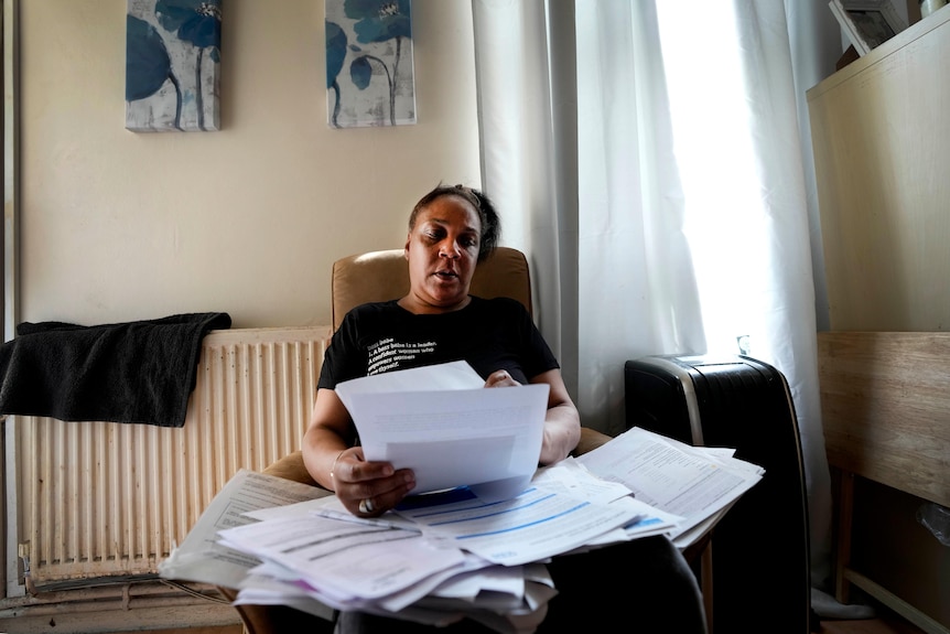 A middle-aged woman sorts through paper at a small table in a plain-looking apartment.