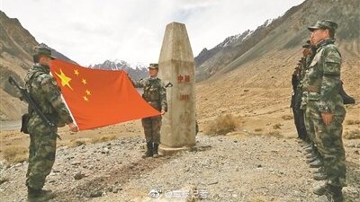 In a brown mountain valley, Chinese military officers in camouflage unfurl a Chinese flag next to a border marker.