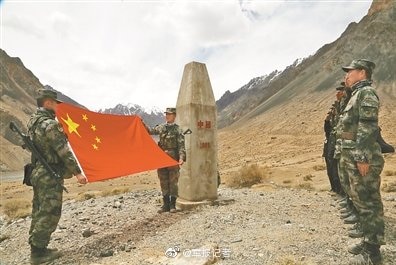 In a brown mountain valley, Chinese military officers in camouflage unfurl a Chinese flag next to a border marker.