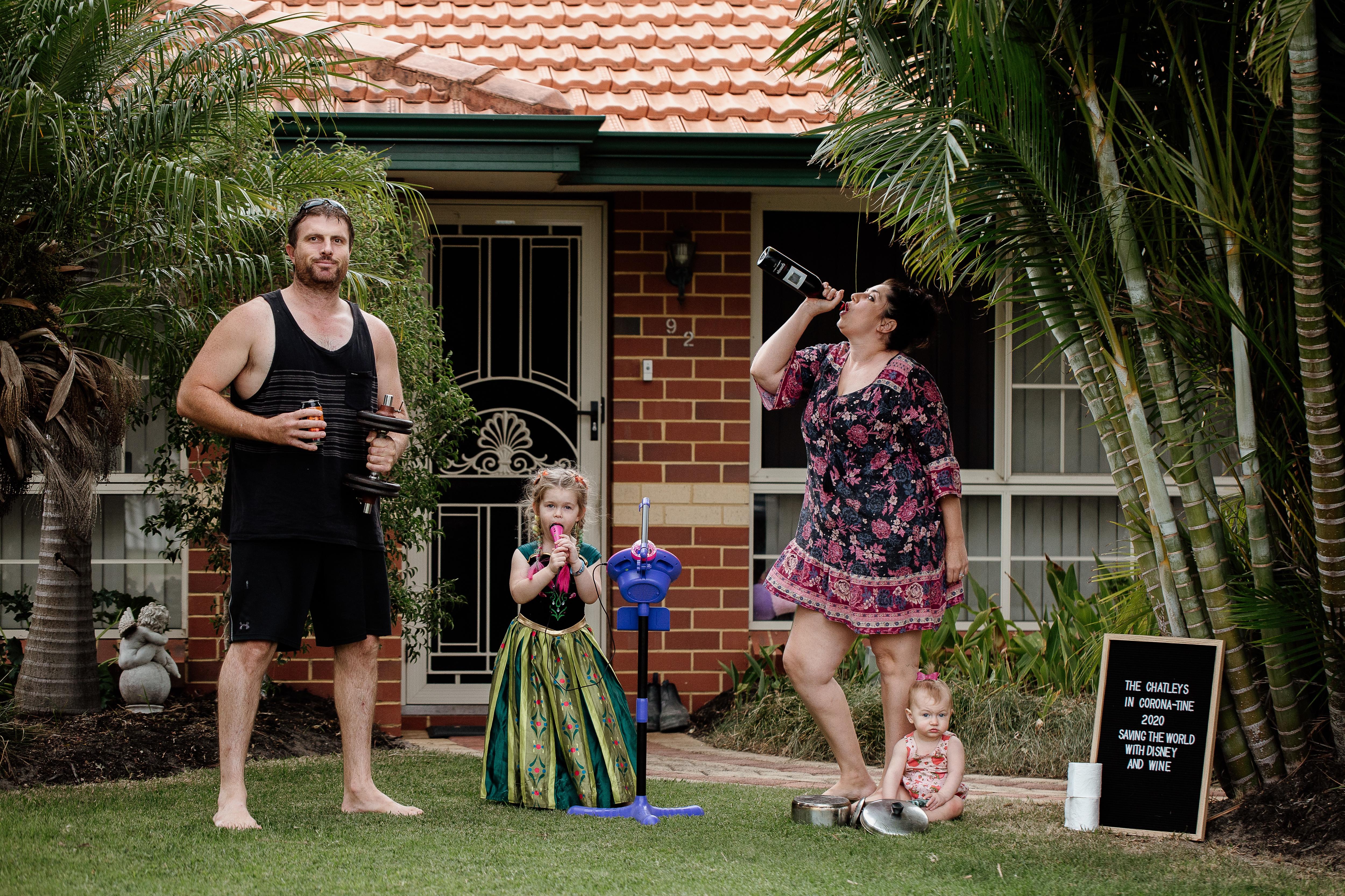 A man stands with weights in hand next to his child in a Disney outfit and his wife who drinks from a wine bottle