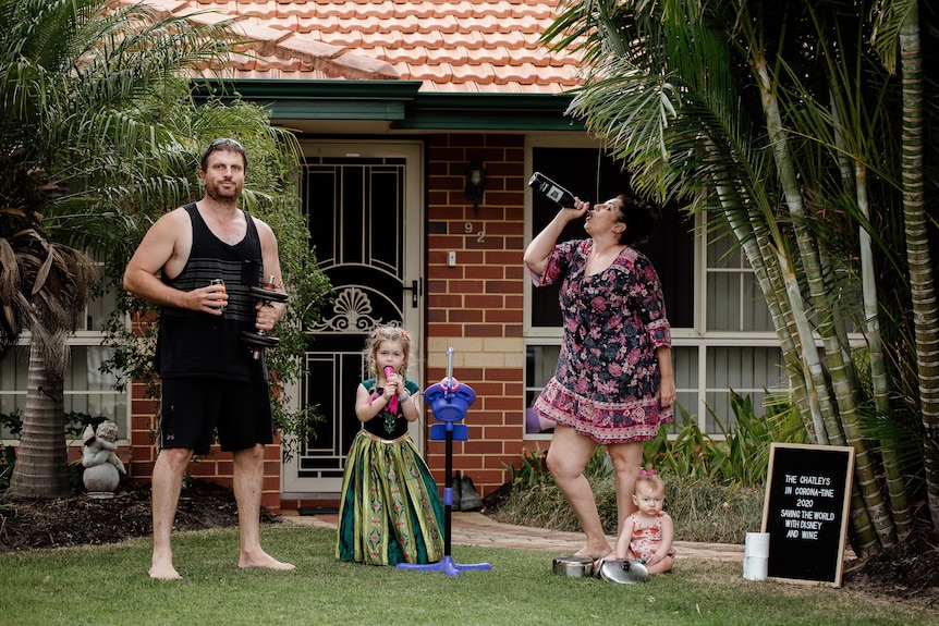 A man stands with weights in hand next to his child in a Disney outfit and his wife who drinks from a wine bottle