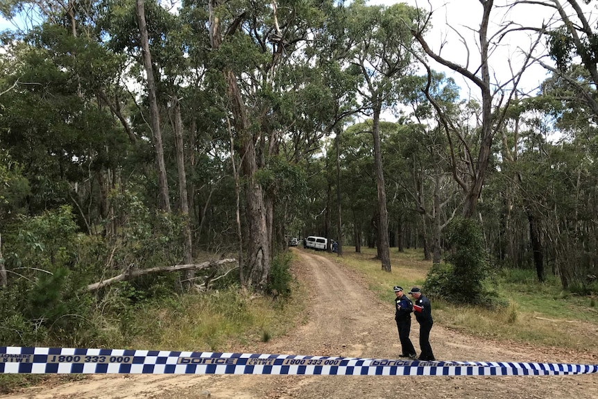 Two police officers stand on a dirt track that leads into dense bushland, blocked by police tape.