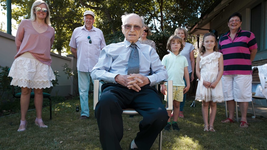 A picture of an elderly man seated in front of his standing family on his 100th birthday.