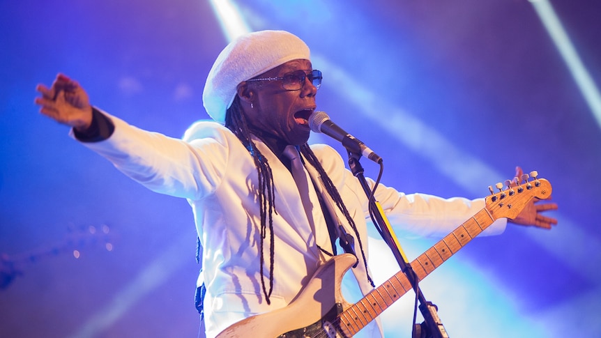 Nile Rodgers stands onstage, bathed in blue light and arms outstretched, wearing a white suit and hat with guitar
