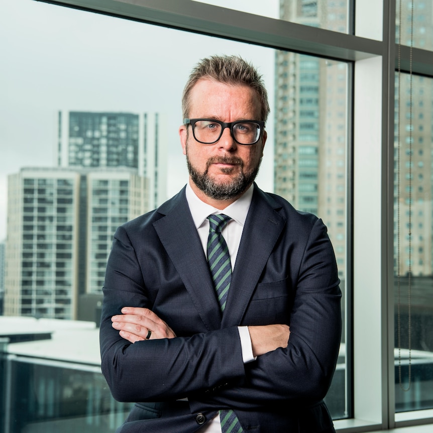 Man wearing glasses with arms crossed looking at camera with city background.