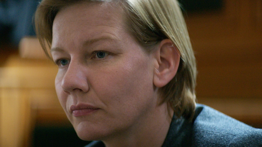 A film still of Sandra Hüller, a middle-aged woman with short blonde hair, in a courtroom. She has a stern expression.