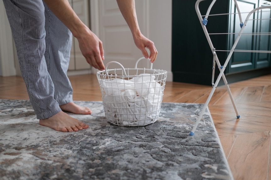 A man in pyjamas bends over to pick up a washing basket filled with white clothes.