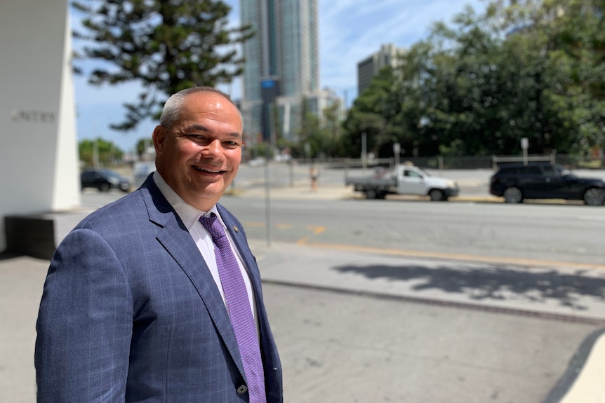 Man in a suit and tie smiling at the camera with a highrise building behind him