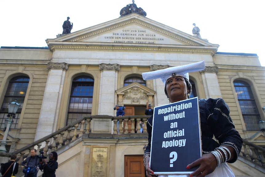 Kobieta z Namibii trzyma znak z napisem "Wracasz do domu bez przeprosin?" Ofiarom ludobójstwa w Namibii. 
