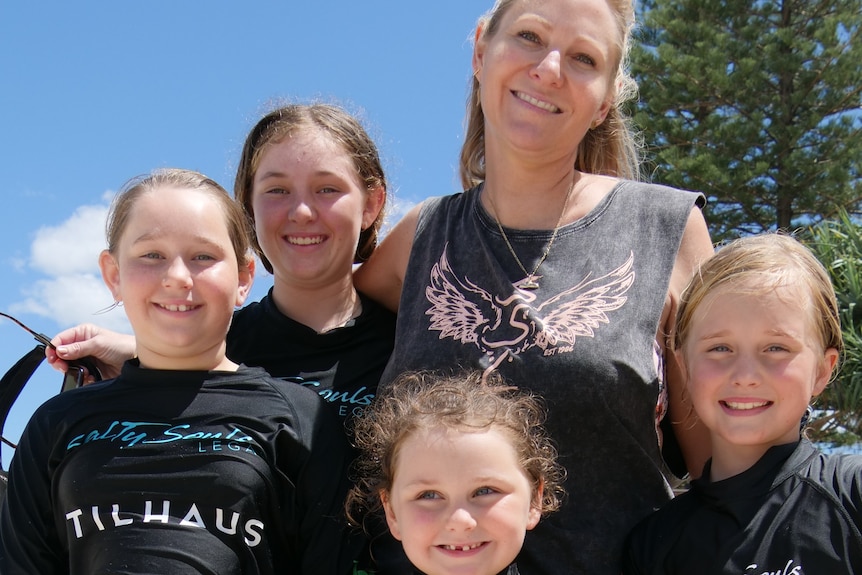 Blonde woman smiling on sunny day at beach with four young girls around her and trees behind