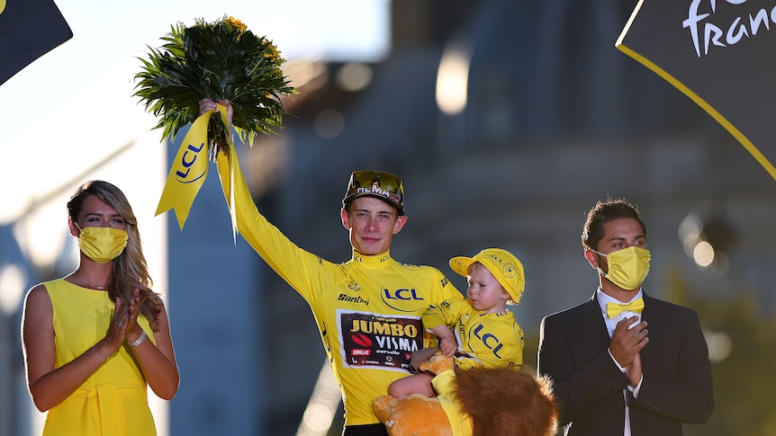 The yellow jersey winner at Tour de France holds his daughter in one arm and a bouquet in the other as he waves to the crowd.