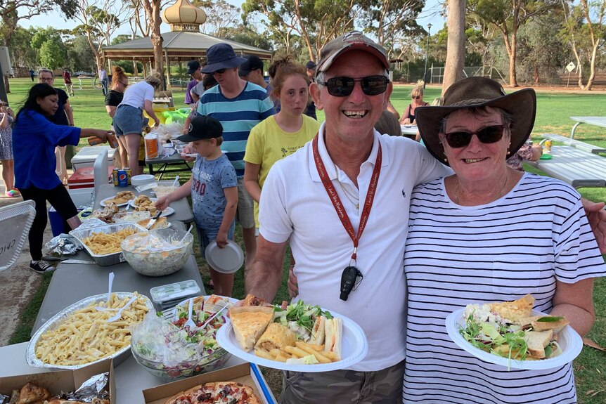 An older male and female couple stand next to a table covered in food in a park, a gazebo and a dozen people stand behind.