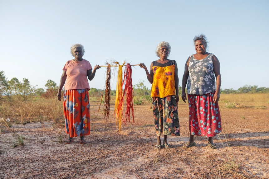 Three Burarra women in their 50s and 60s wear colourful shirts and skirts and hold freshly dyed pandanus thread.
