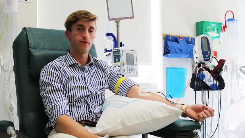 Ben Henry, dressed in a shirt and pants, sits on a hospital chair as blood is drawn from a needle in his left forearm.