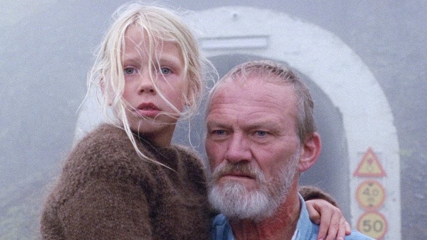 An older man covered in blood, holding a young girl standing in front of a tunnel on a foggy day in the film A White, White Day