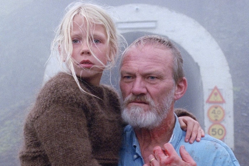 An older man covered in blood, holding a young girl standing in front of a tunnel on a foggy day in the film A White, White Day