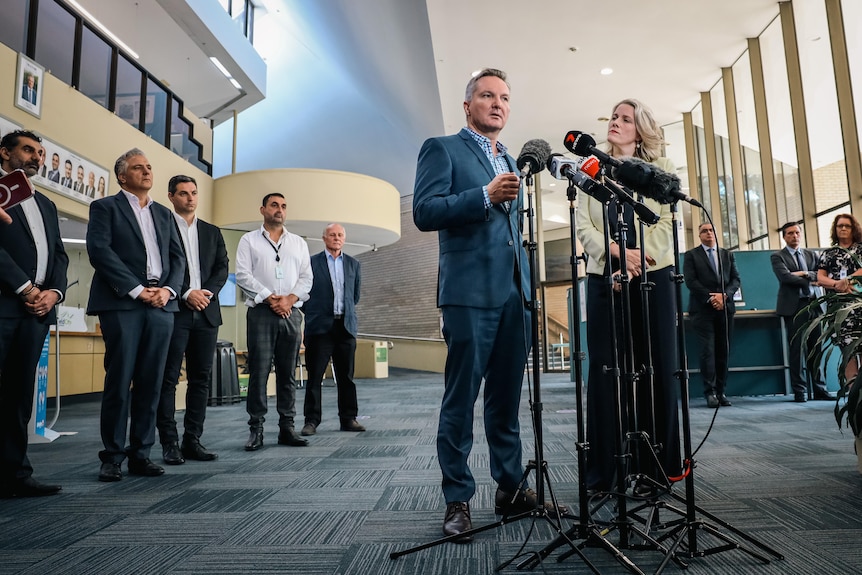 A man and a woman talk to microphones while a line of men stand to their side