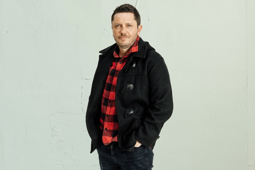 A man with short dark hair poses for a photo, with his hands in his jean pockets. He wears a black jacket over a red plaid shirt