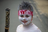 An Indigenous boy wearing white body paint and a red headband stands smiling and holding a didgeridoo.