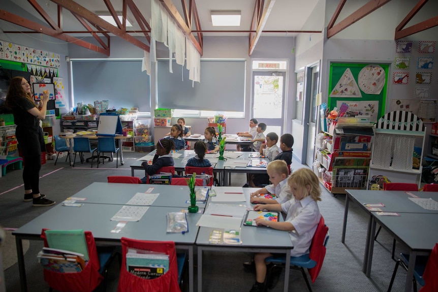 A class room of 12 grade prep children talk amongst themselves as the teacher helps one student