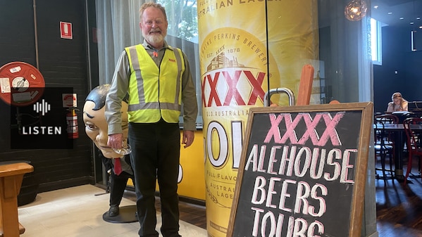 A man in a high-vis jacket has a jolly smile as he stands next to a jumbo quadruple-X sign. . Has Audio.