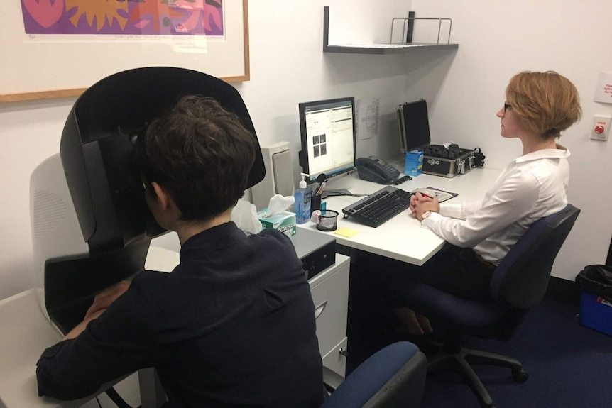 A woman sits with her head resting in a white metal box with a researcher next to her.