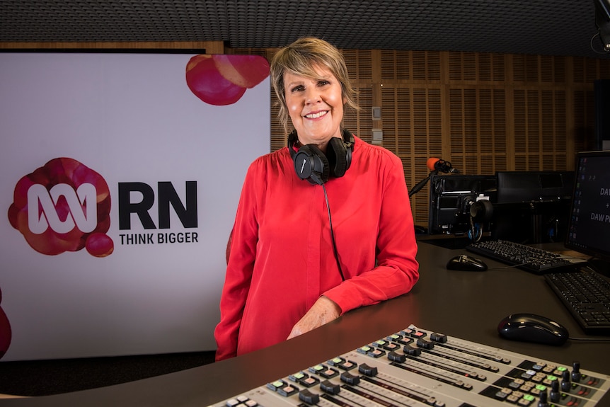 Woman with headphones around neck leaning against radio studio desk.