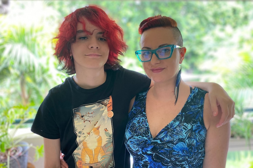 A teenager in a black t-shirt and dyed red hair stands next to a woman in a blue dress, green glasses and dyed red hair.