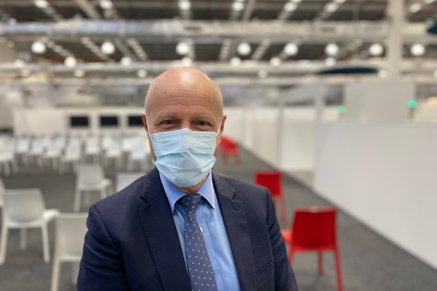 A man in a suit wearing a surgical mask in a room filled with plastic chairs.