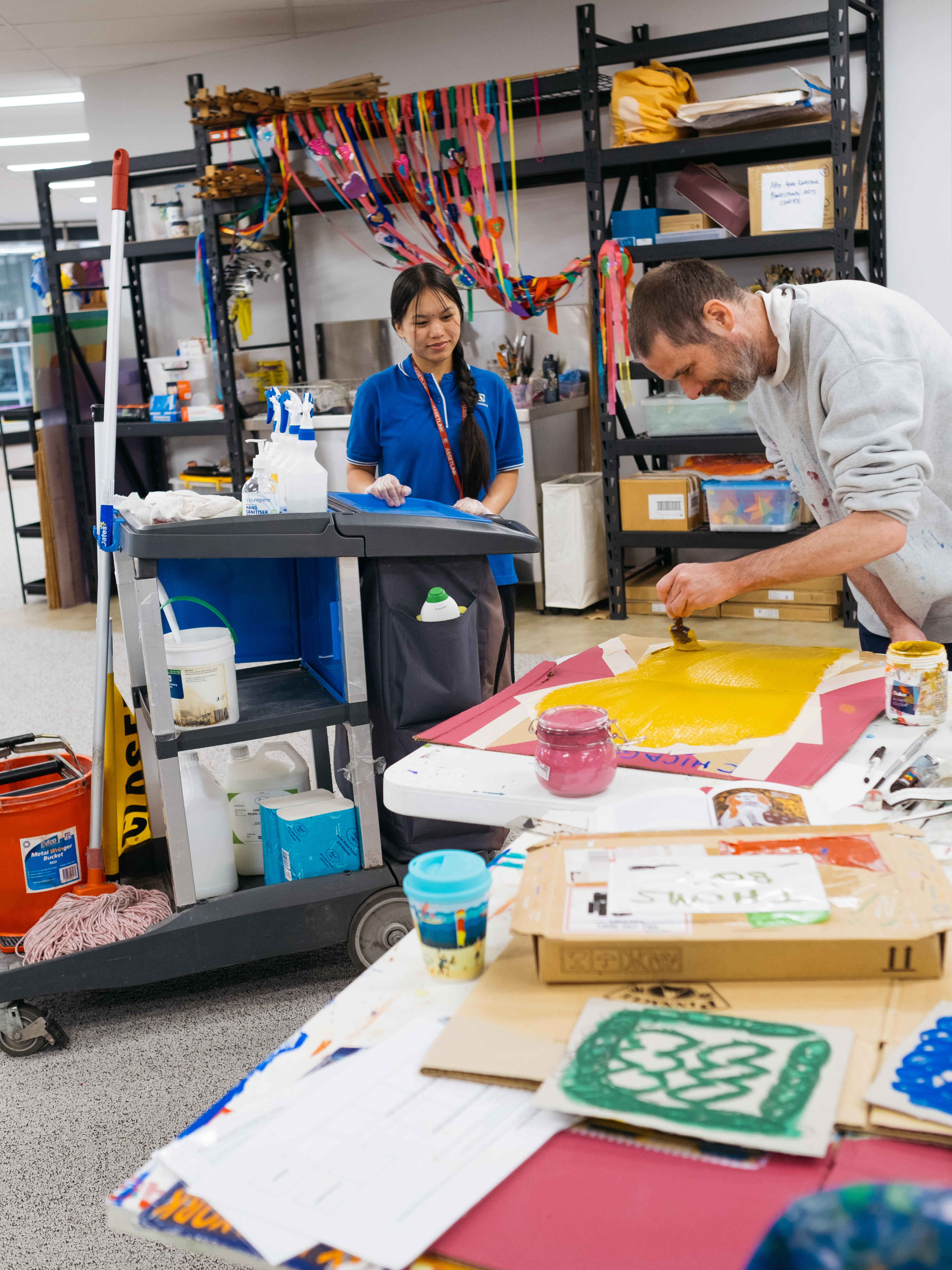 Cleaner Subita, with long dark hair and a blue t-shirt, stands with her cleaning trolley watching artist Thom paint at his bench