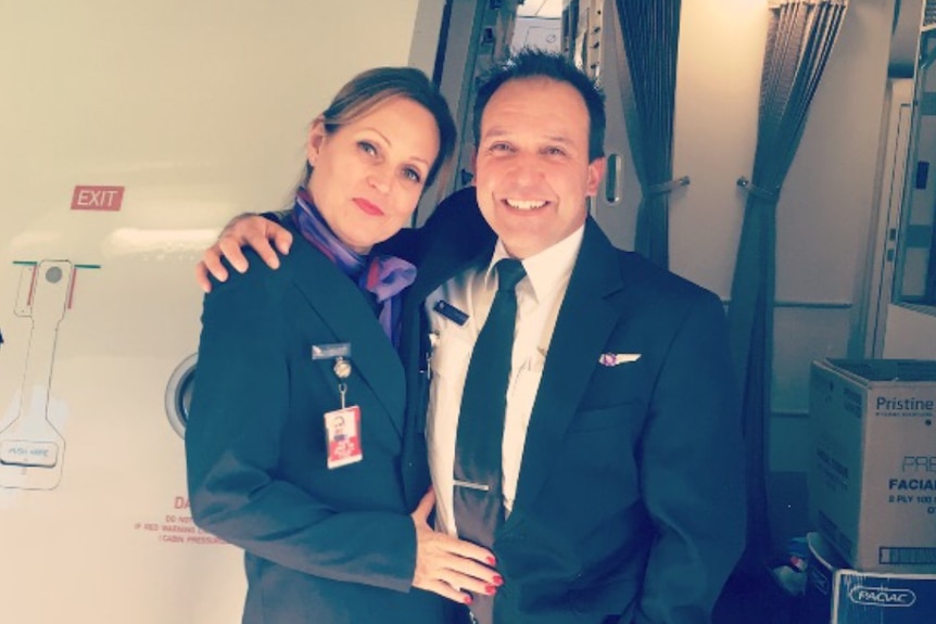 A pilot in uniform stands with his partner, a flight stewardess, outside an aircraft door.