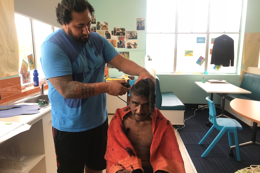 A boy sits in a chair getting his hair cut short by a man with hair clippers.