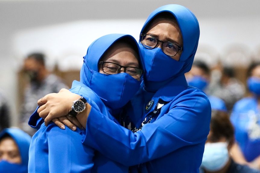 Two women in blue face masks and veils cry while hugging each other