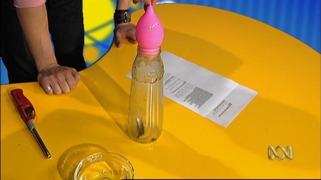 Person stands beside table with a bottle full of water and a ballon-type object wedged in the neck