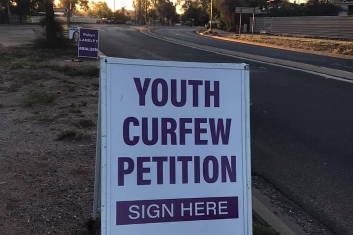 A sandwich board sign reading "Youth curfew petition, sign here" standing on the side of a suburban road