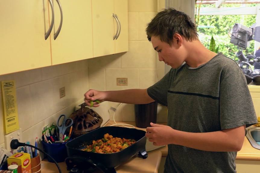 A 12-year-old boy in a grey shirt cooking a meal in a small kitchen.