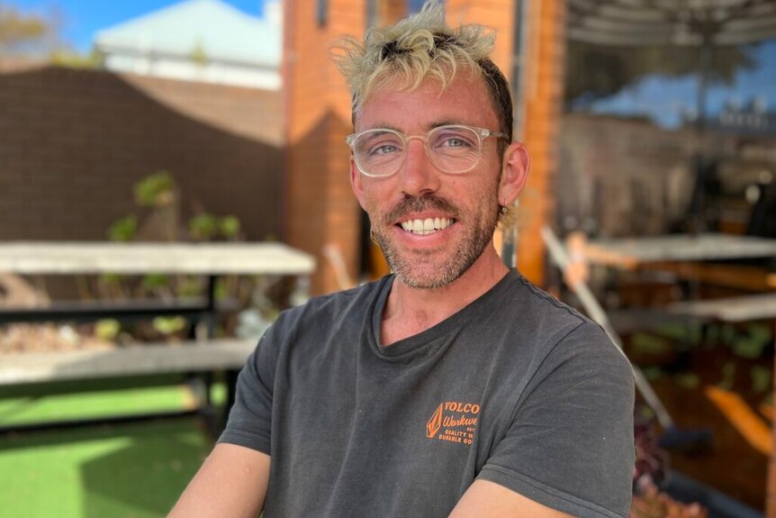 A smiling man with dyed curly blond hair and spectacles, sitting outside on a sunny day.