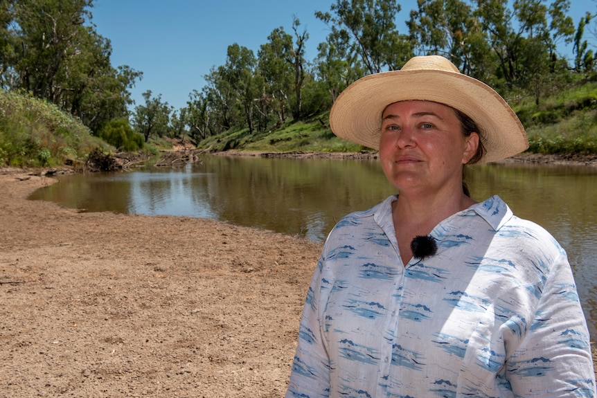 A woman in a white shirt and yellow hat standing beside a river. 