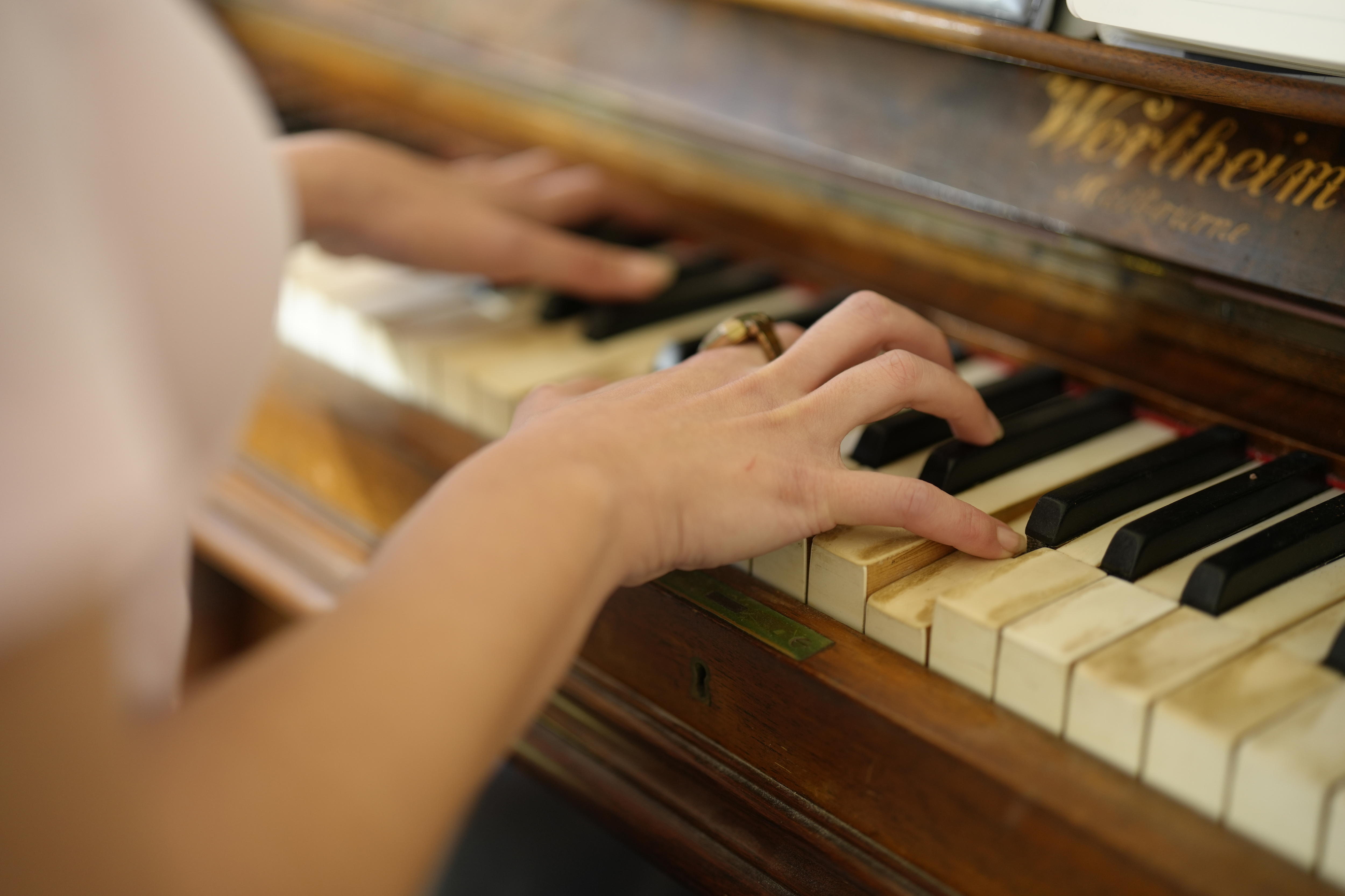 Two hands play a brown, wooden piano. One hand is wearing a gold ring.