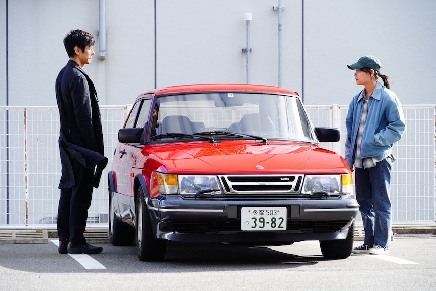 Japanese man with dark hair and clothing stands looking at Japanese woman with teal cap and denim jacket; a red car between them