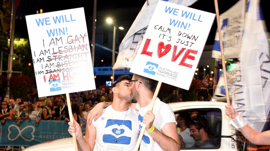 Two men kiss while holding a sign which says 'Calm down, it's just love.'