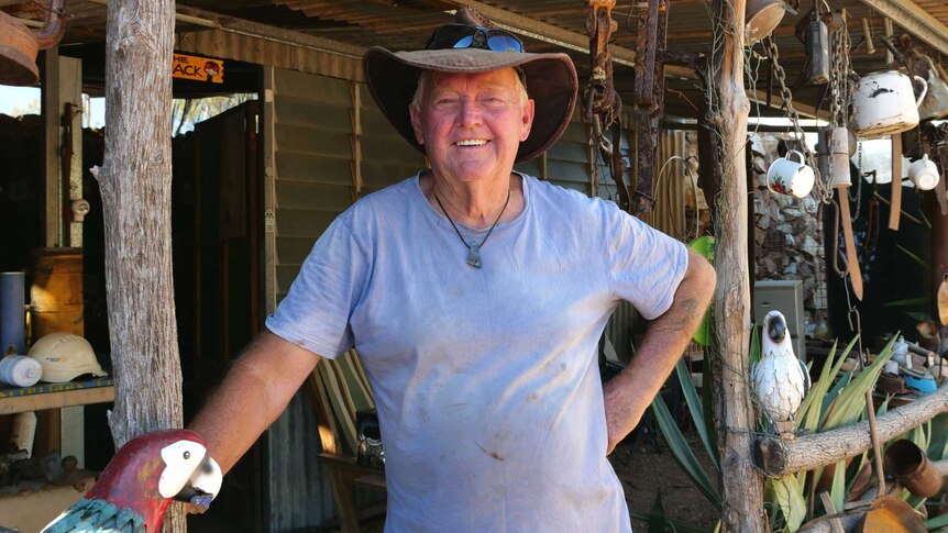 A man looking directly at the camera, standing in front of a tin shed with lots of hanging objects. He is smiling.