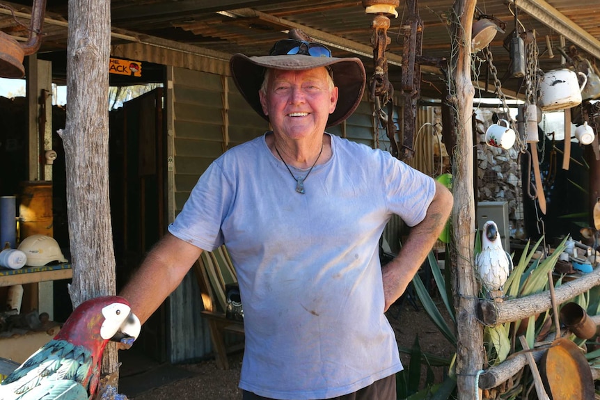 A man looking directly at the camera, standing in front of a tin shed with lots of hanging objects. He is smiling.