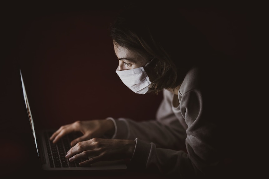 A light-skinned person wearing a facemask uses a laptop in a dark room
